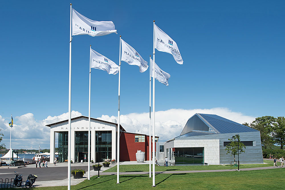 Tre flaggstänger med hissade Marinmuseumflaggor, gräs och på lite håll Marinmuseum sedd från entrén.