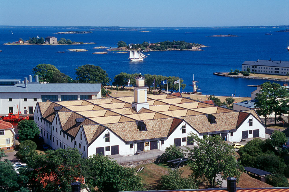 Slup- och barkasskjulet med sitt karaktärristiska tegeltak. I bakgrunden vatten, skärgård och en skuta för fulla segel.