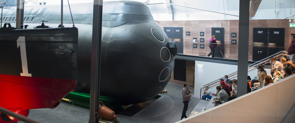 Man står framför en grupp ungdomar som sitter på en läktare. Strax bakom ligger ubåtarna HMS Neptun och HMS Hajen. Bilden är tagen inuti Marinmuseums ubåtshall och delar av utställningen i två plan finns med på bilden. 
