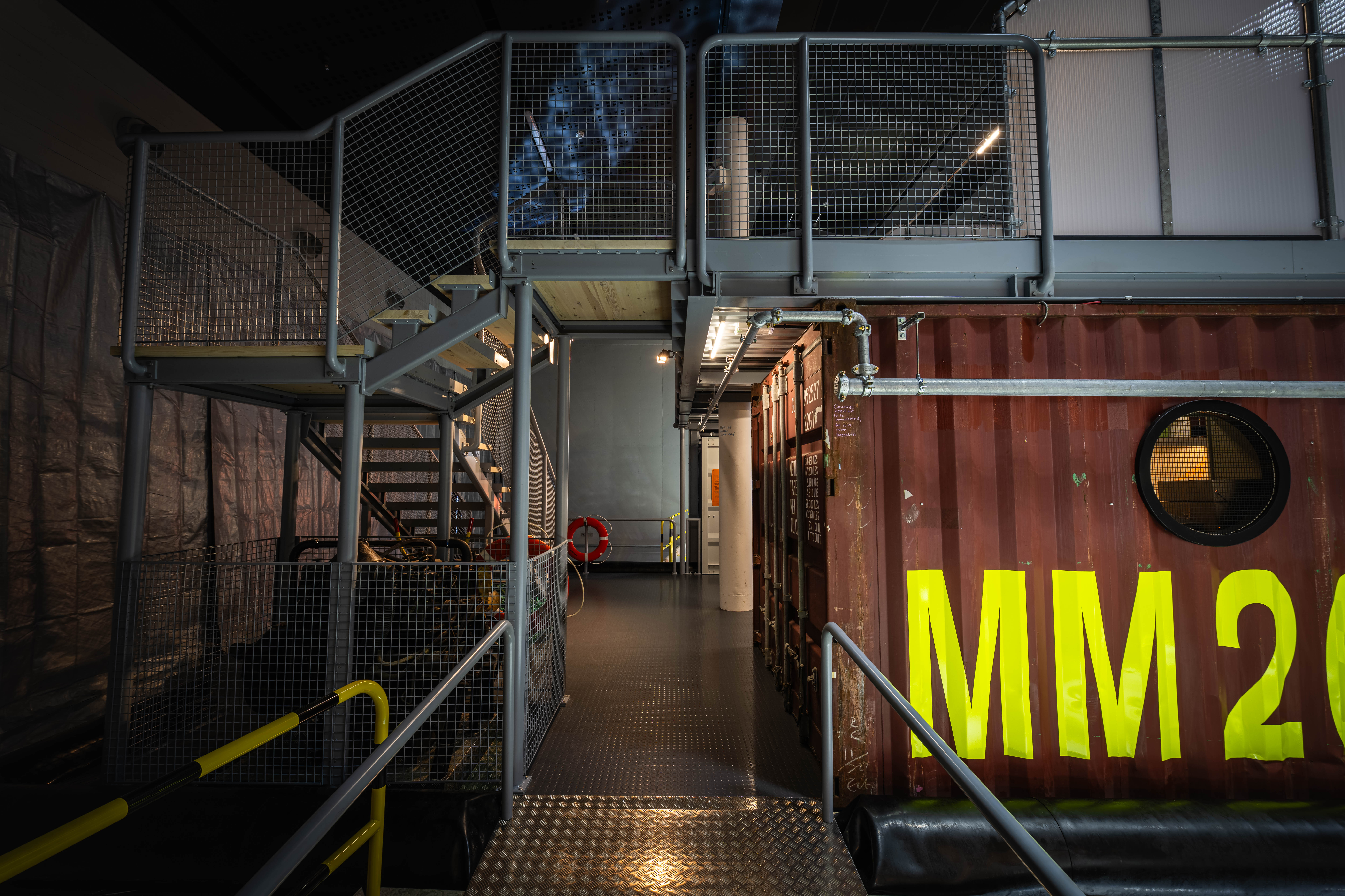 Industriell museimiljö med en trappa och gångbro i metall bredvid en fraktcontainer med stora gula bokstäver.