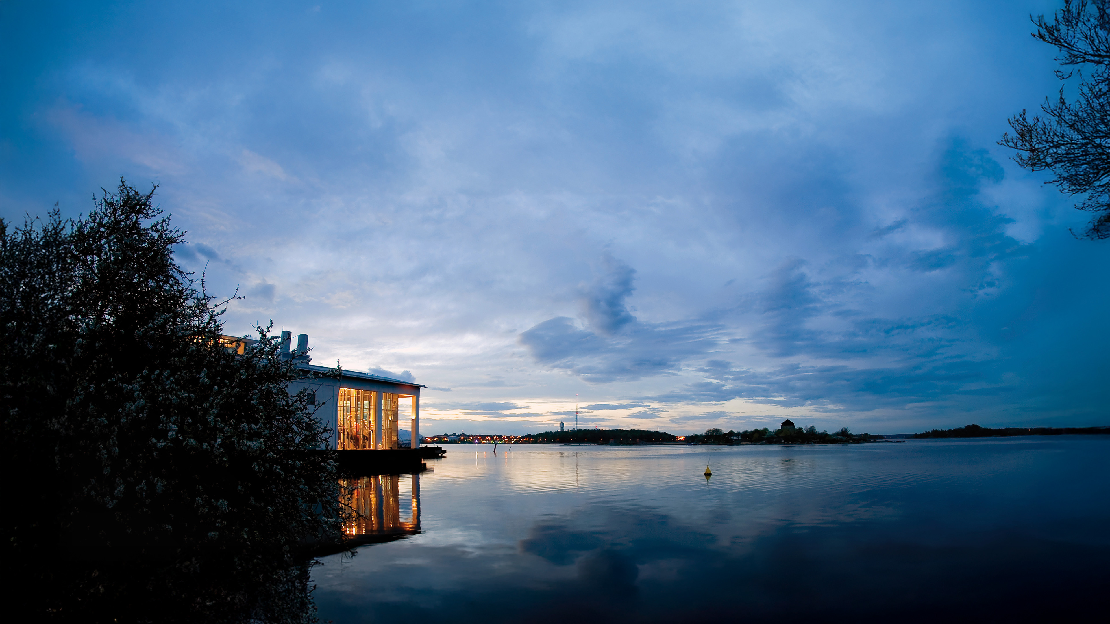 Marinmuseum i Karlskrona vid vattnet i skymning, med byggnaden upplyst inifrån och himlen speglande sig i det lugna havet.