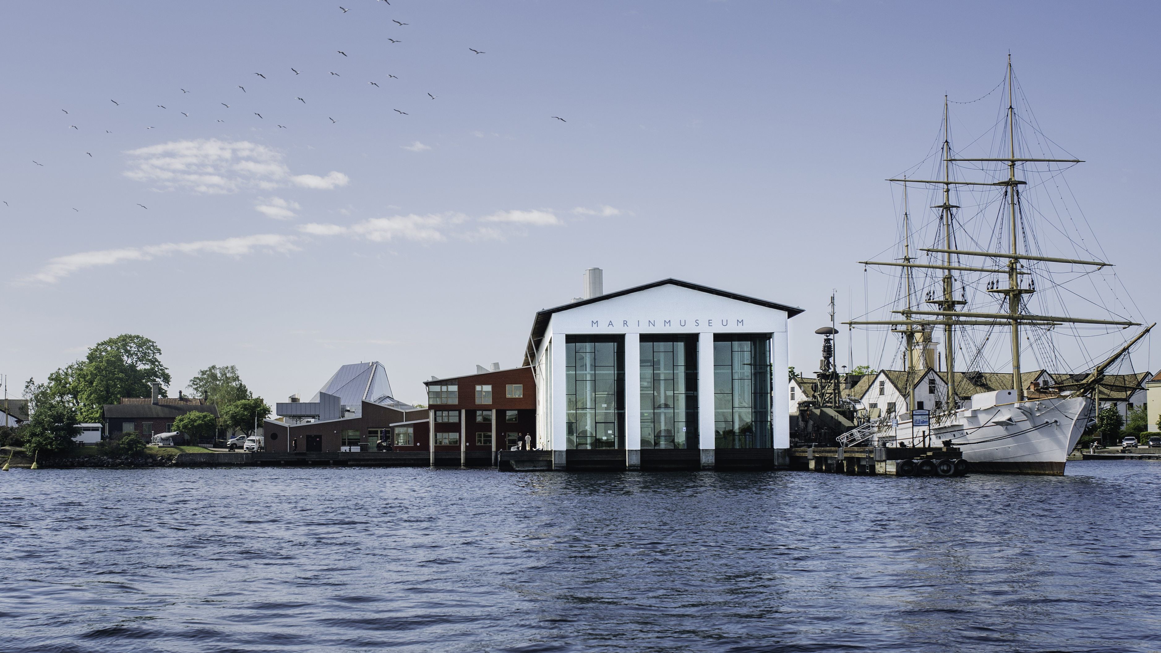 The Naval Museum in Karlskrona seen from the water on a sunny day, with a sailing ship moored by the museum and birds flying in the sky.