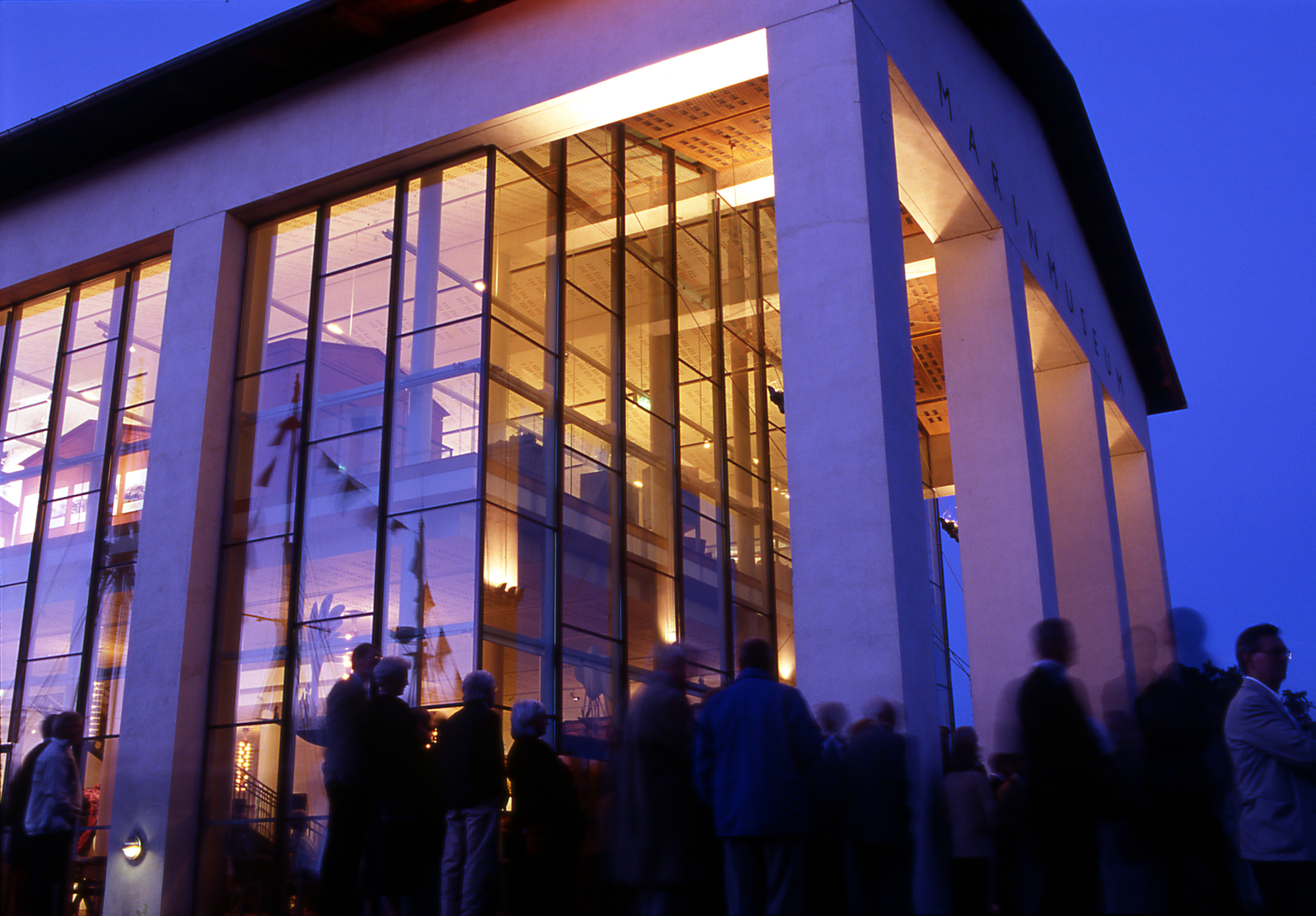 Kvällsbild av Marinmuseums entré i Karlskrona, där människor samlas utanför den upplysta glasfasaden under en blå skymningshimmel.