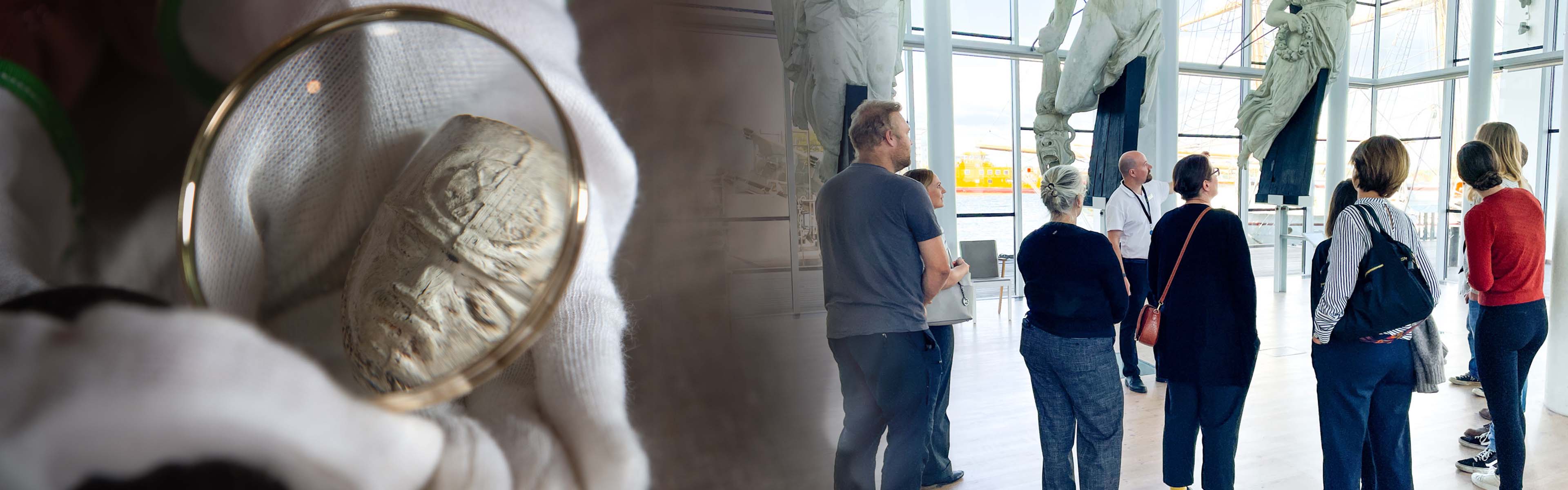 A group of people listens to a guide during a museum tour, surrounded by large historic ship figureheads in a bright room with large windows. On the left, a close-up shows a gloved hand holding a carved object under a magnifying glass.