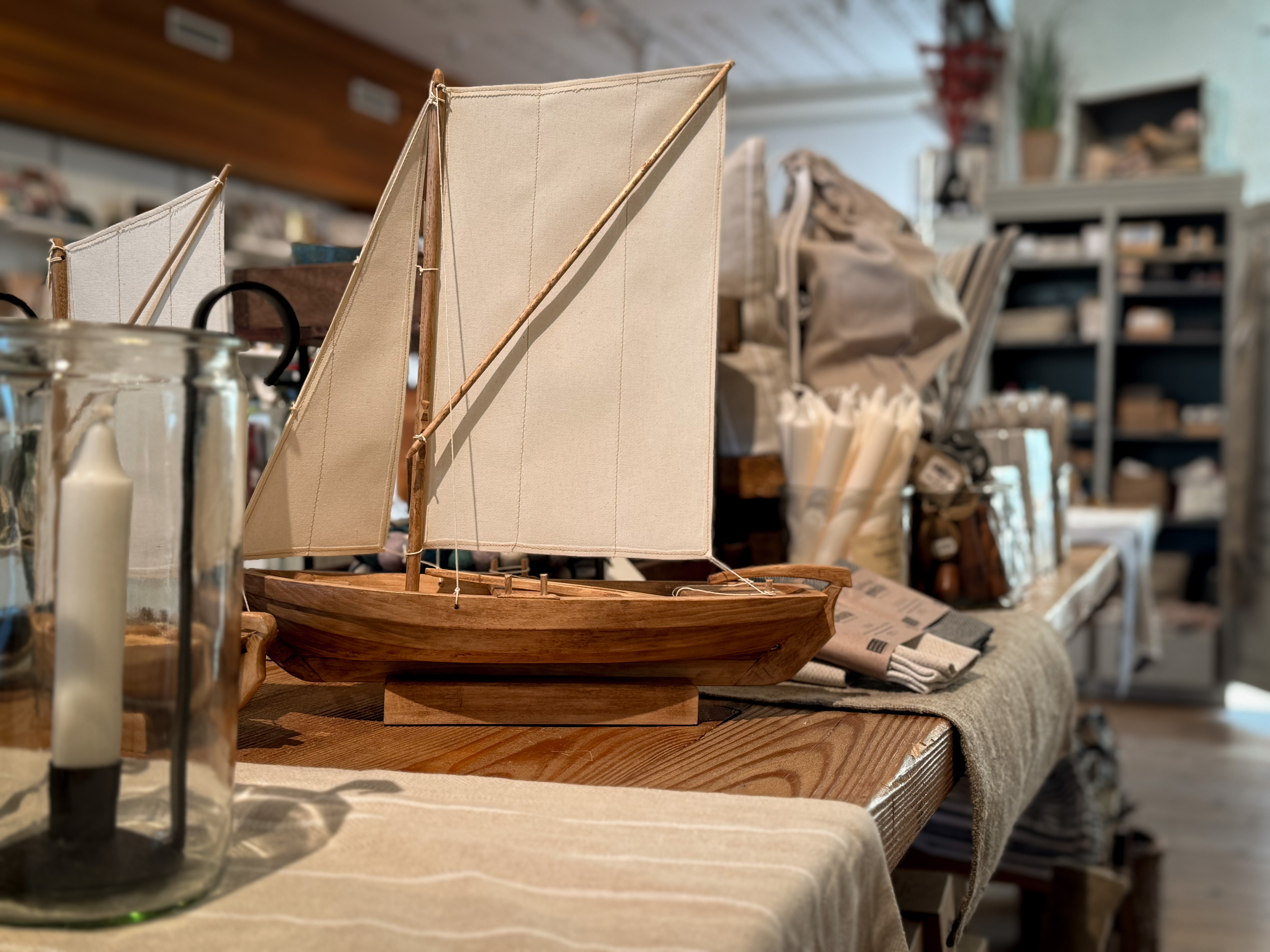 Trämodell av ett segelfartyg med vita segel på ett bord i museibutiken på Marinmuseum, omgiven av maritima produkter och inredningsdetaljer.