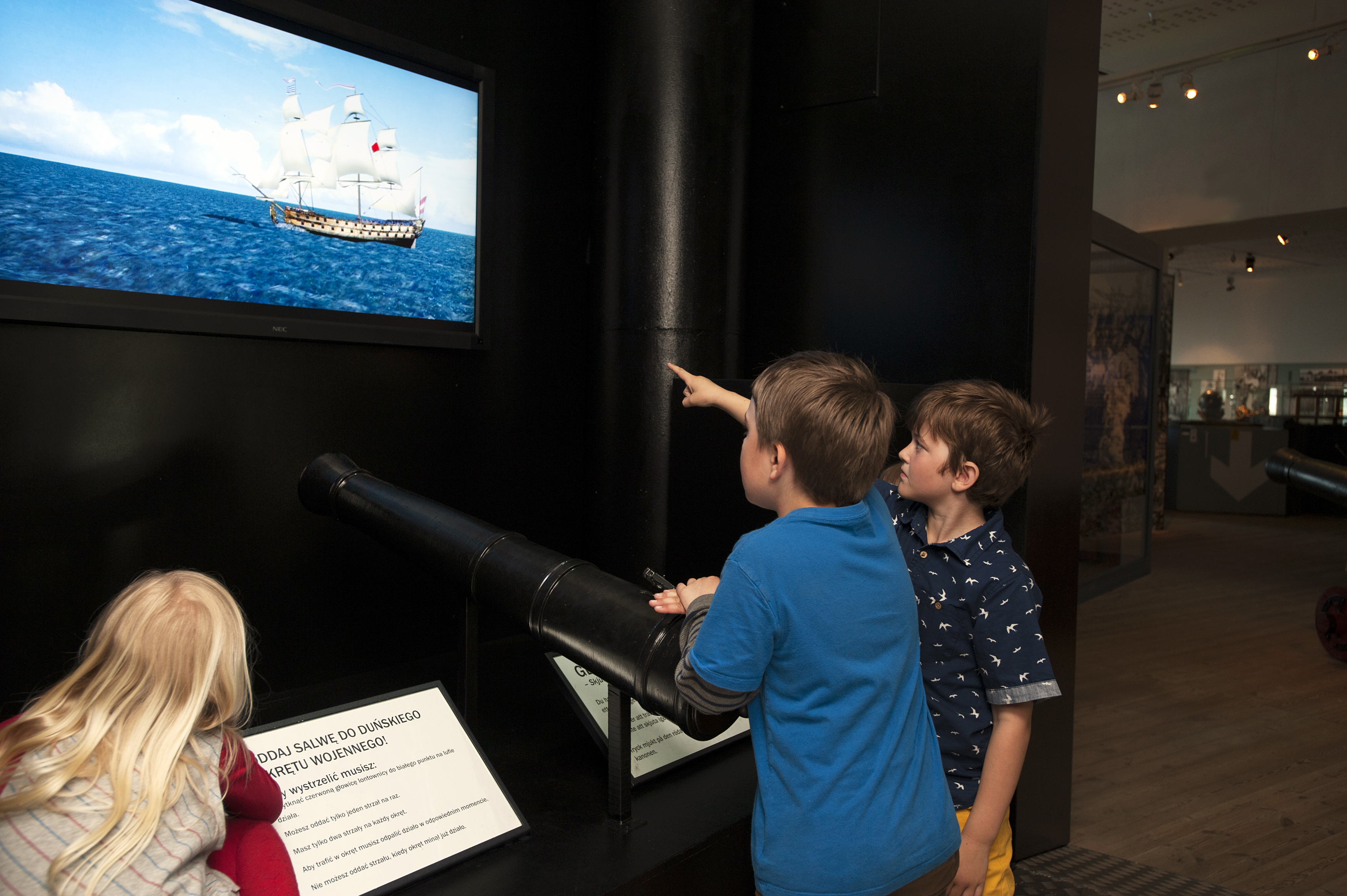 Tre barn deltar i en interaktiv aktivitet på Marinmuseum där de siktar med en kanon mot ett fartyg på en skärm.