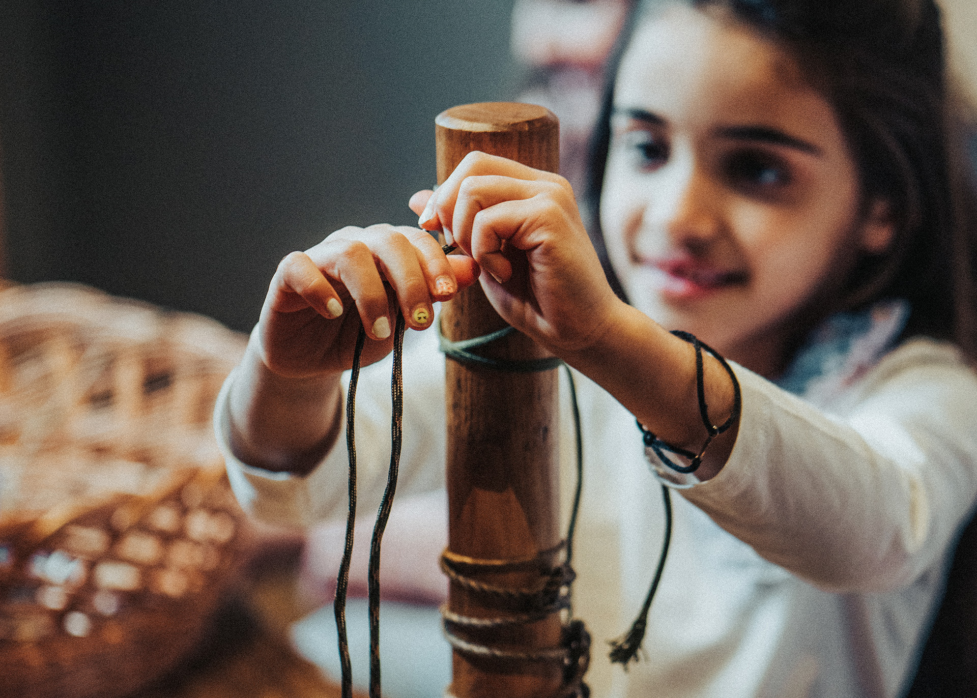 A child tying strings around a wooden pole. The child has painted nails and is wearing a bracelet, looking focused while working with the strings.