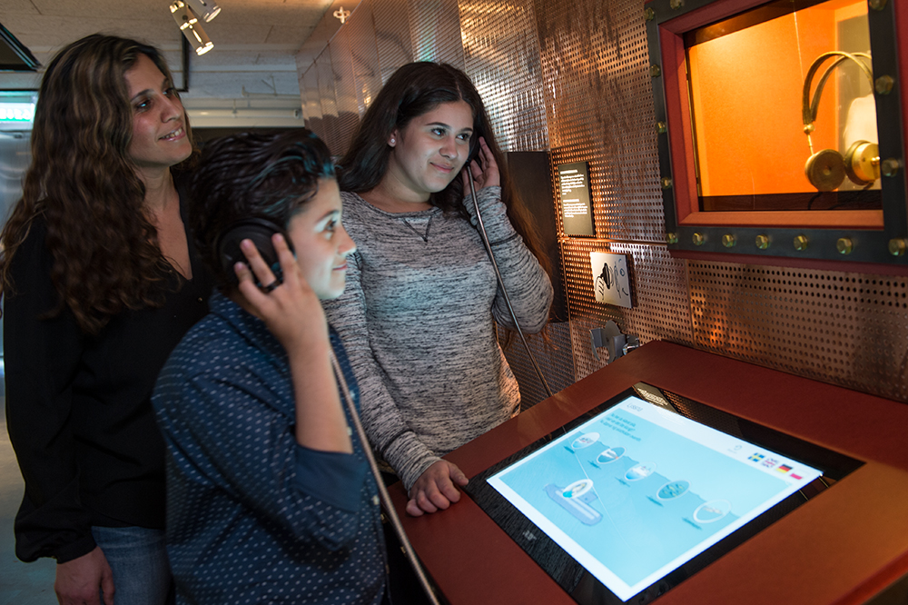 En vuxen och två barn deltar i en interaktiv aktivitet på Marinmuseum där de lyssnar i hörlurar vid en digital station med skärm.