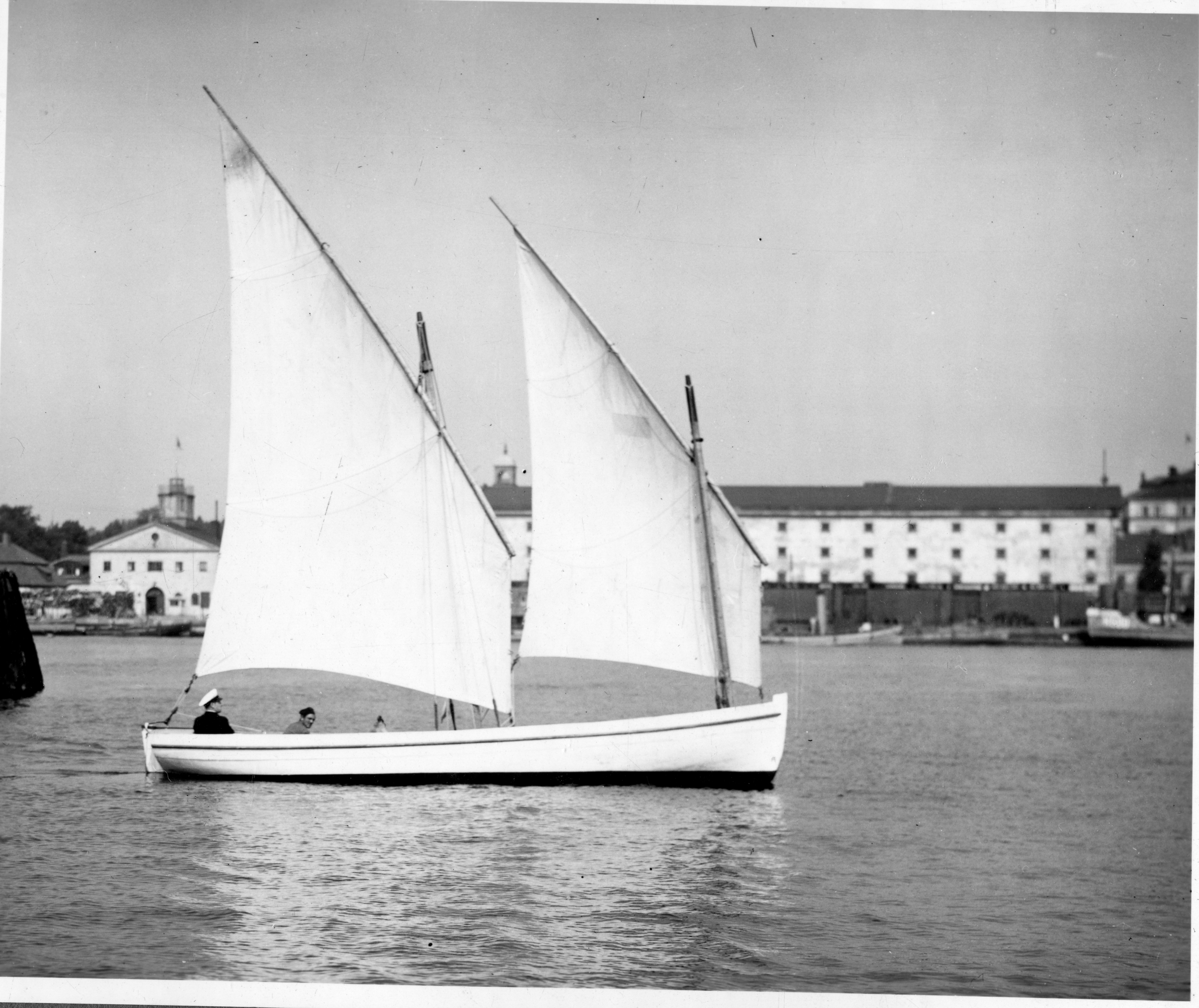 En äldre svartvit bild av en segelbåt med två segel och två personer ombord på lugnt vatten.