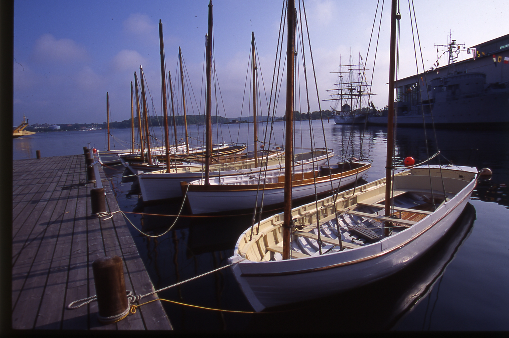 Flera träbåtar ligger förtöjda vid en brygga i stilla vatten med större fartyg i bakgrunden.