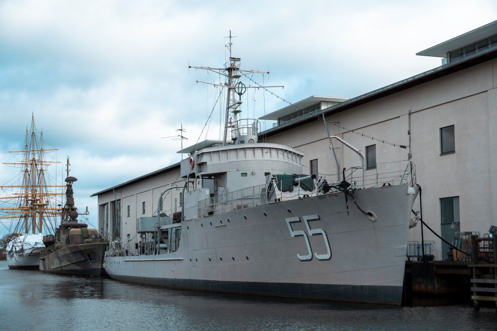 Large gray ship is moored in front of white building. On the side of the ship are the numbers "55".