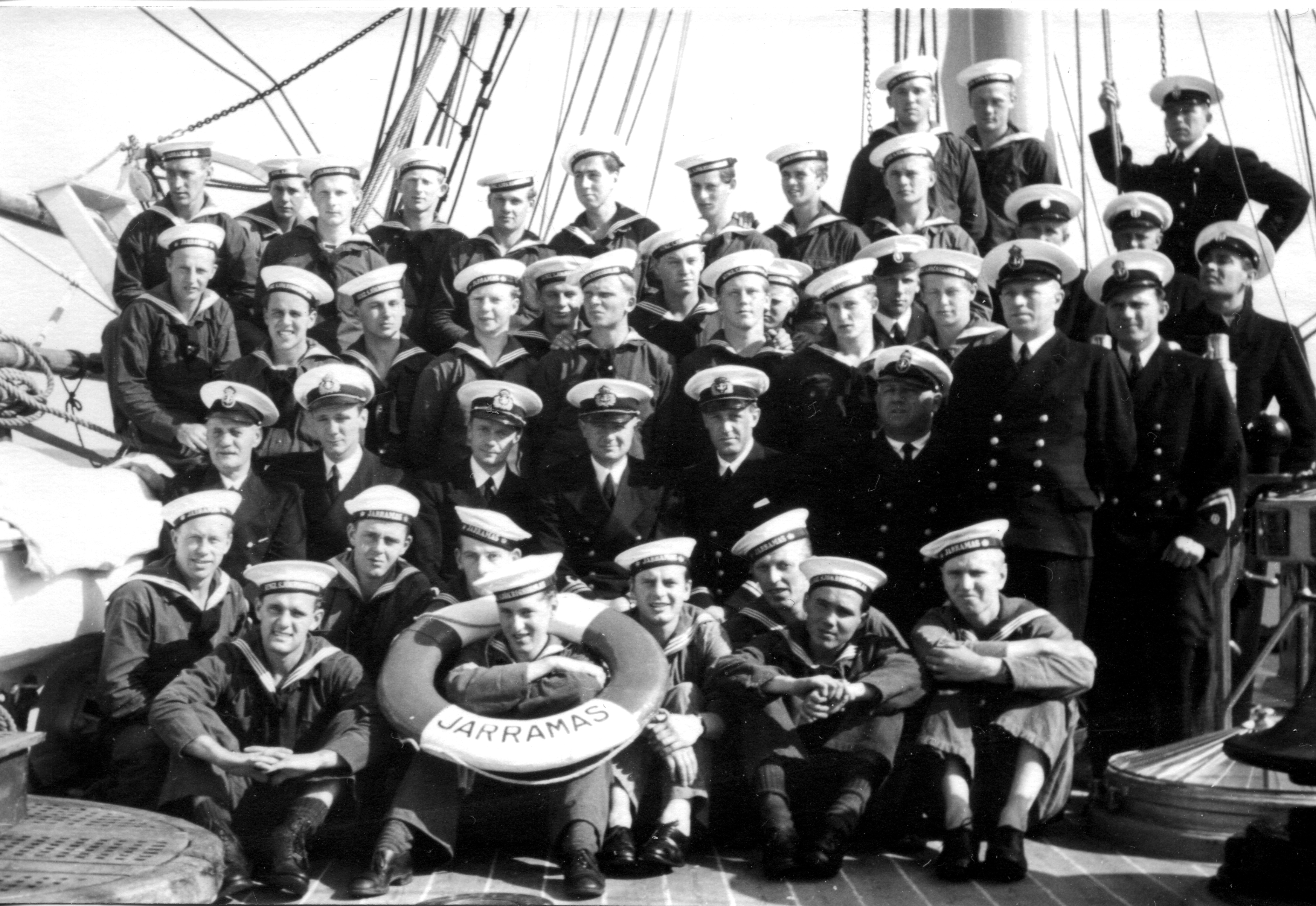 Gruppfoto av sjömän och officerare på däck av fartyget "Jarramas". De bär uniformer och flera sitter med en livboj märkt ”JARRAMAS” i förgrunden.