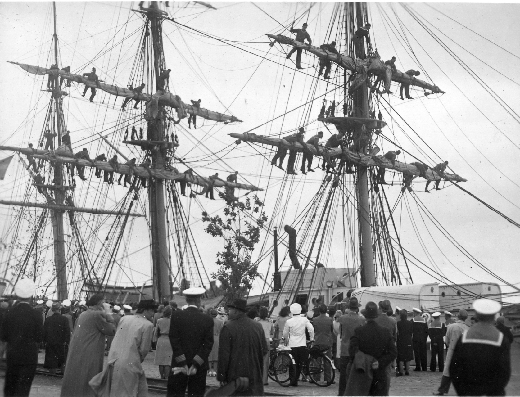 Sjömän klättrar och balanserar på rårna högt upp i riggen på ett segelfartyg medan en folksamling tittar på från kajen nedanför.