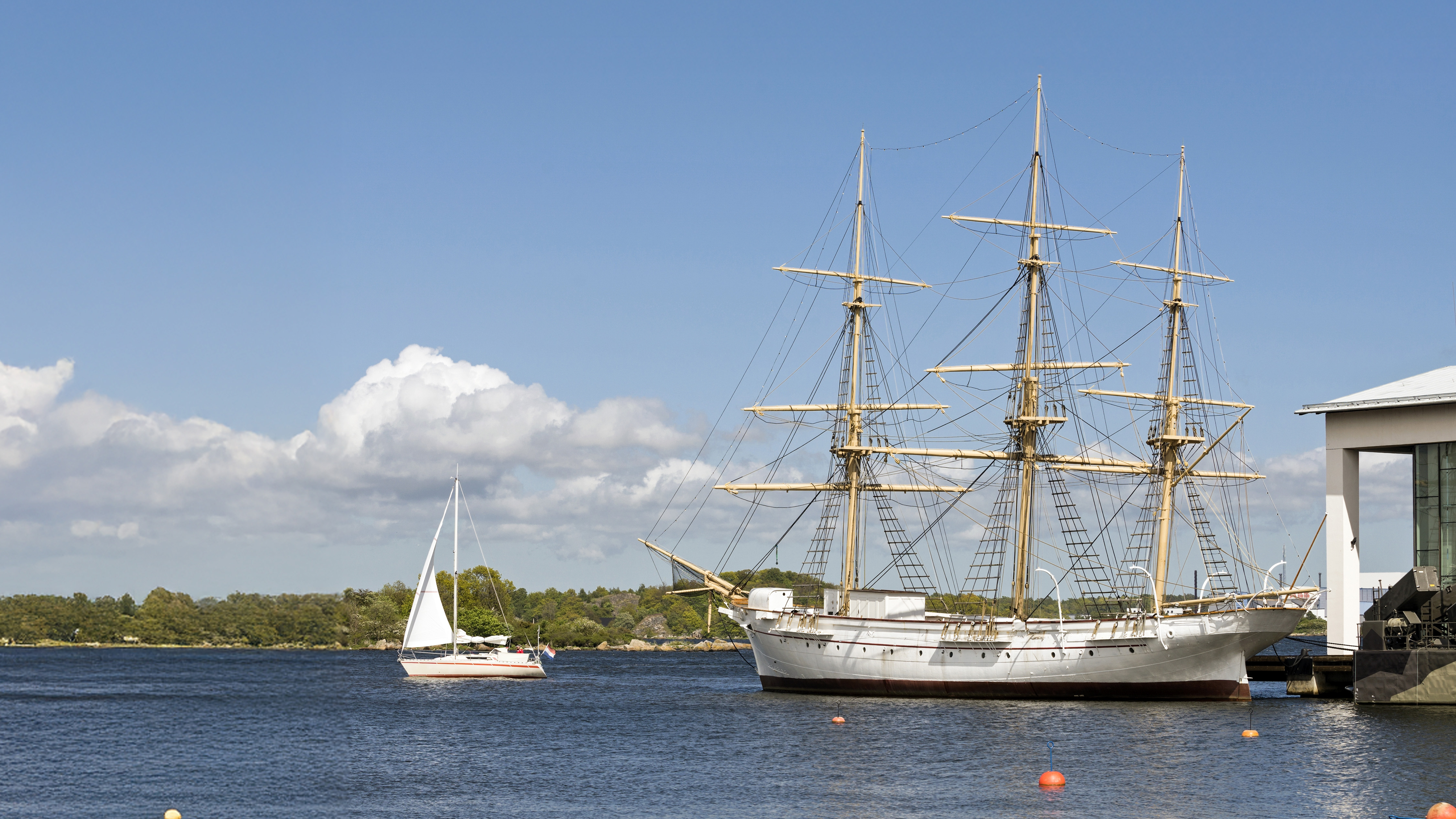 The full-rigged ship Jarramas moored at the Naval Museum in Karlskrona on a sunny day. In the background, a sailboat passes on the calm water under a clear blue sky.