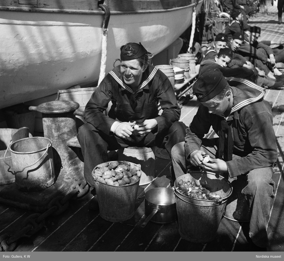 Två sjömän i uniform sitter på ett fartygsdäck och skalar potatis. De har hinkar med potatis framför sig och ser avslappnade ut. Fler besättningsmän syns i bakgrunden.
