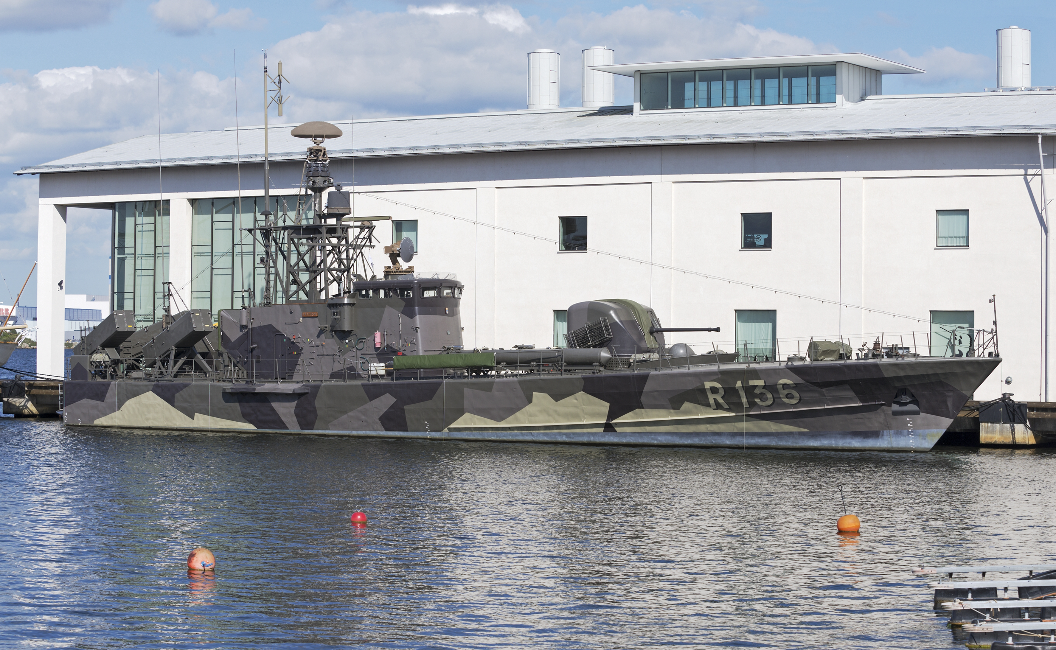 A camouflaged ship is moored next to a large white museum building.