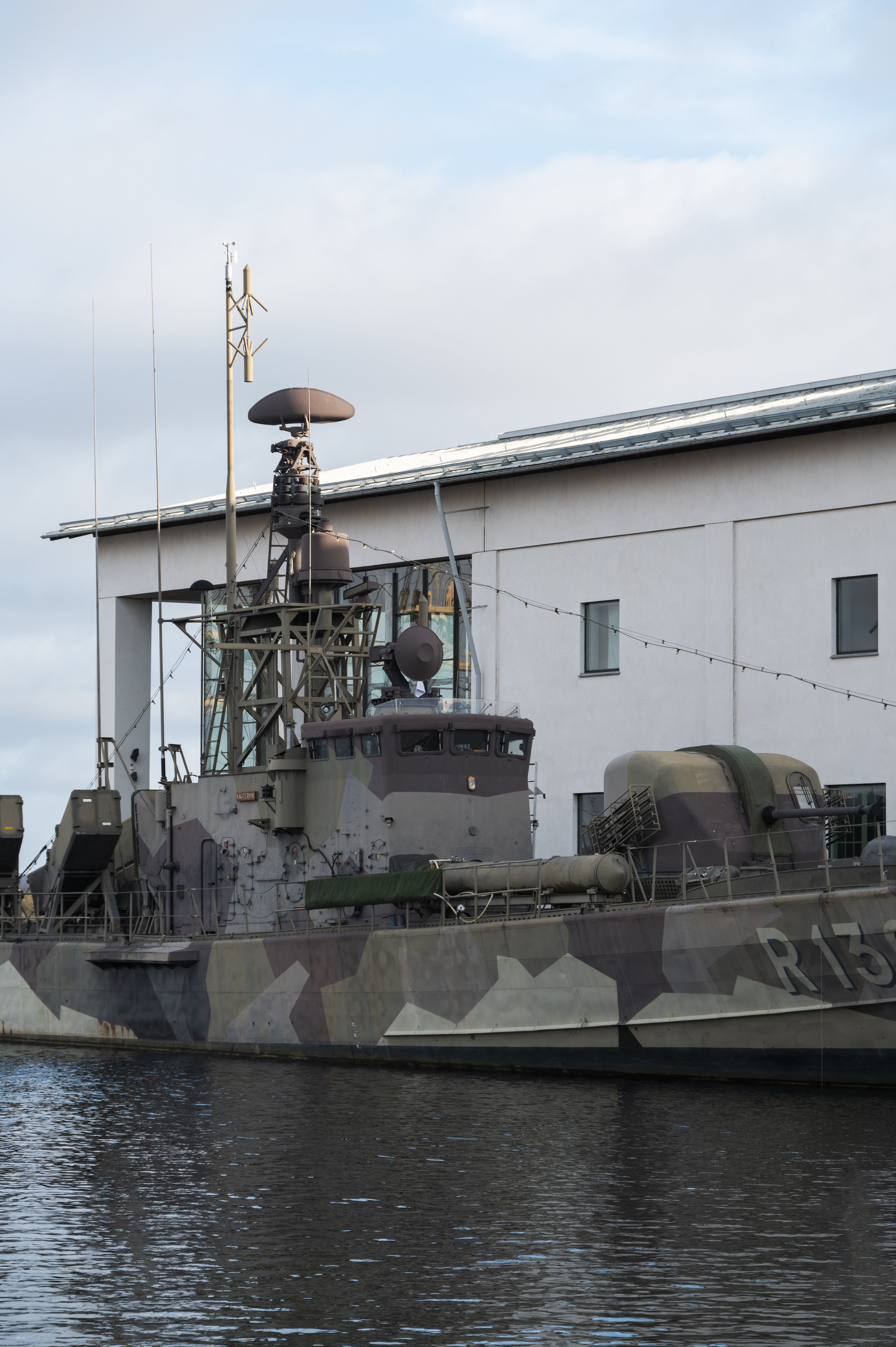 Military vessels with camouflage colors are docked by the white building of the naval museum.