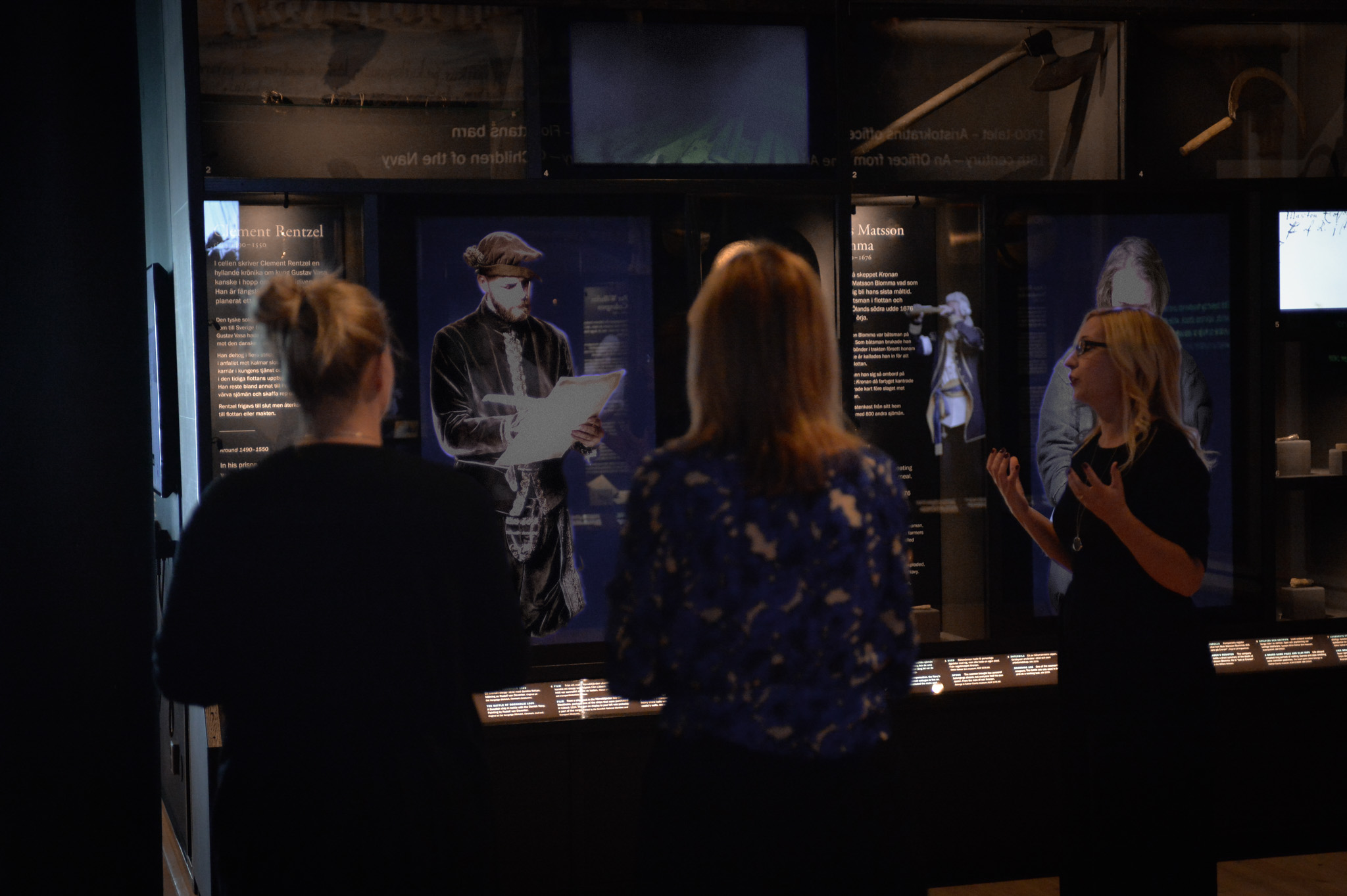 En guide berättar för två besökare i en mörk utställning på Marinmuseum, framför belysta montrar med historiska figurer och texter.