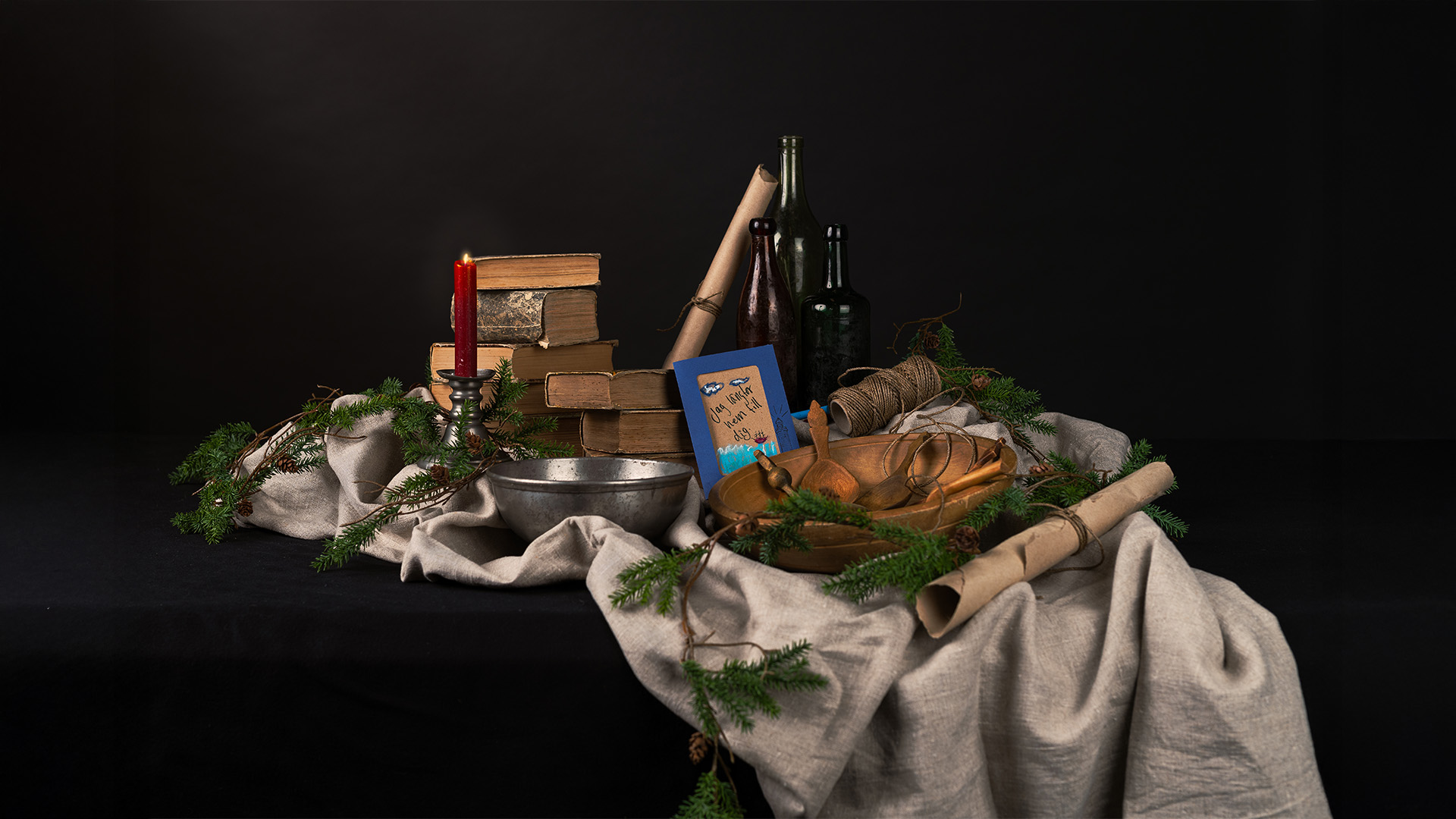 Juldekoration med böcker, ett rött ljus, flaskor, trälådor, ett julkort och grenar, allt på ett tyg.