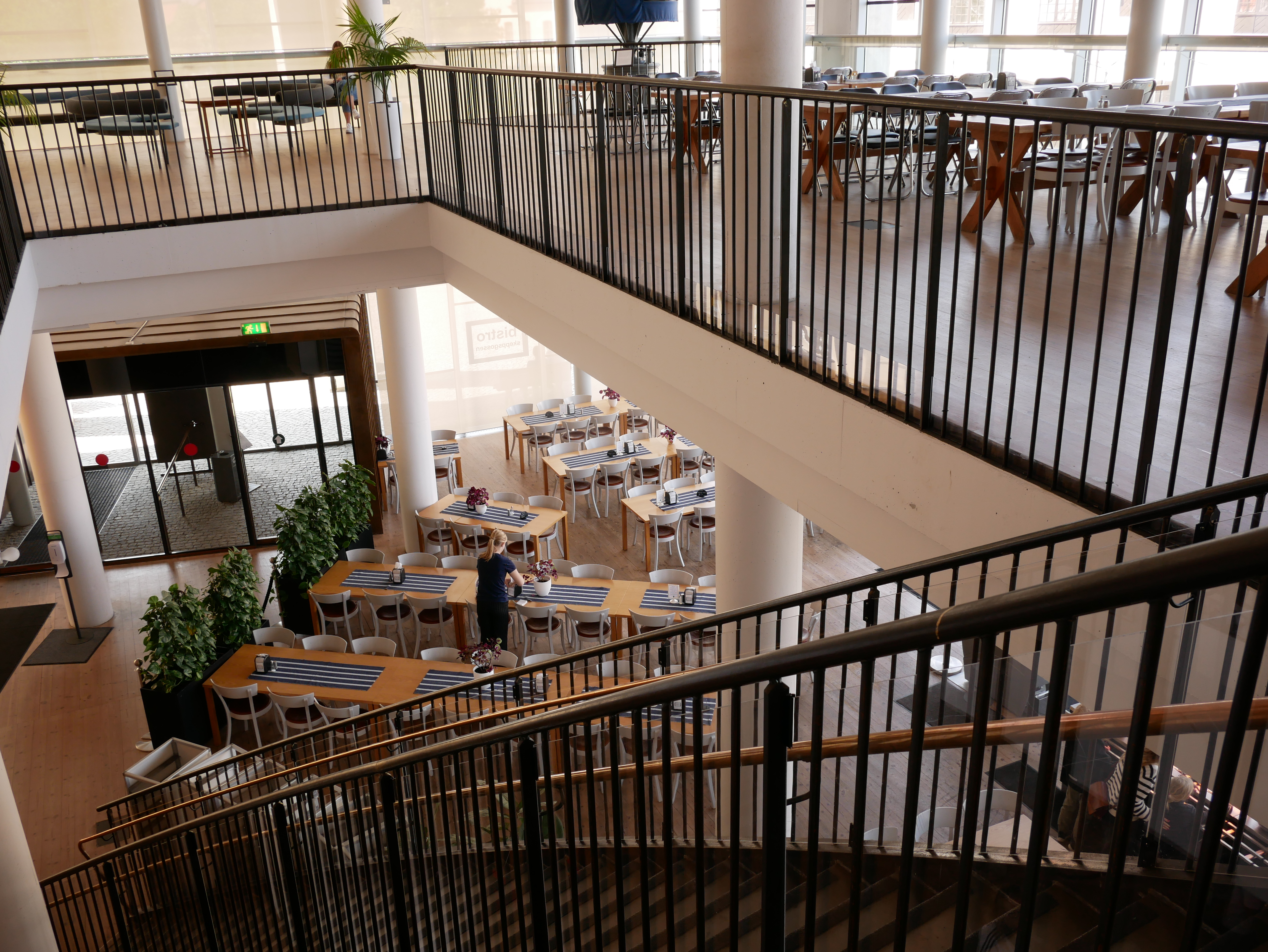 Restaurangen på Marinmuseum med stora fönster, två våningar och dukade bord. En person förbereder matsalen inför servering.