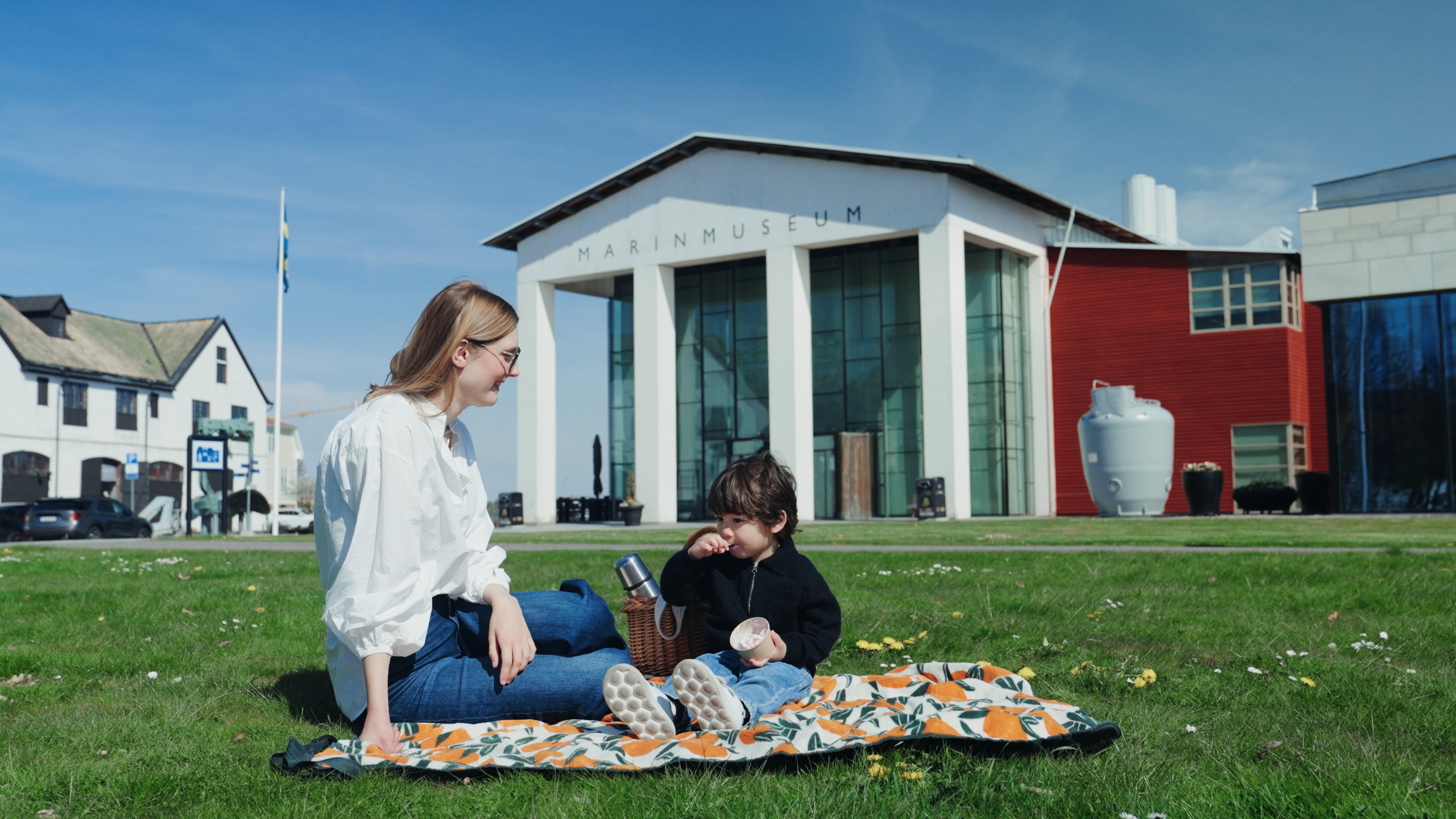 An adult and a child sit on a blanket having a picnic on the lawn outside the Naval Museum in Karlskrona on a sunny day with a clear blue sky.
