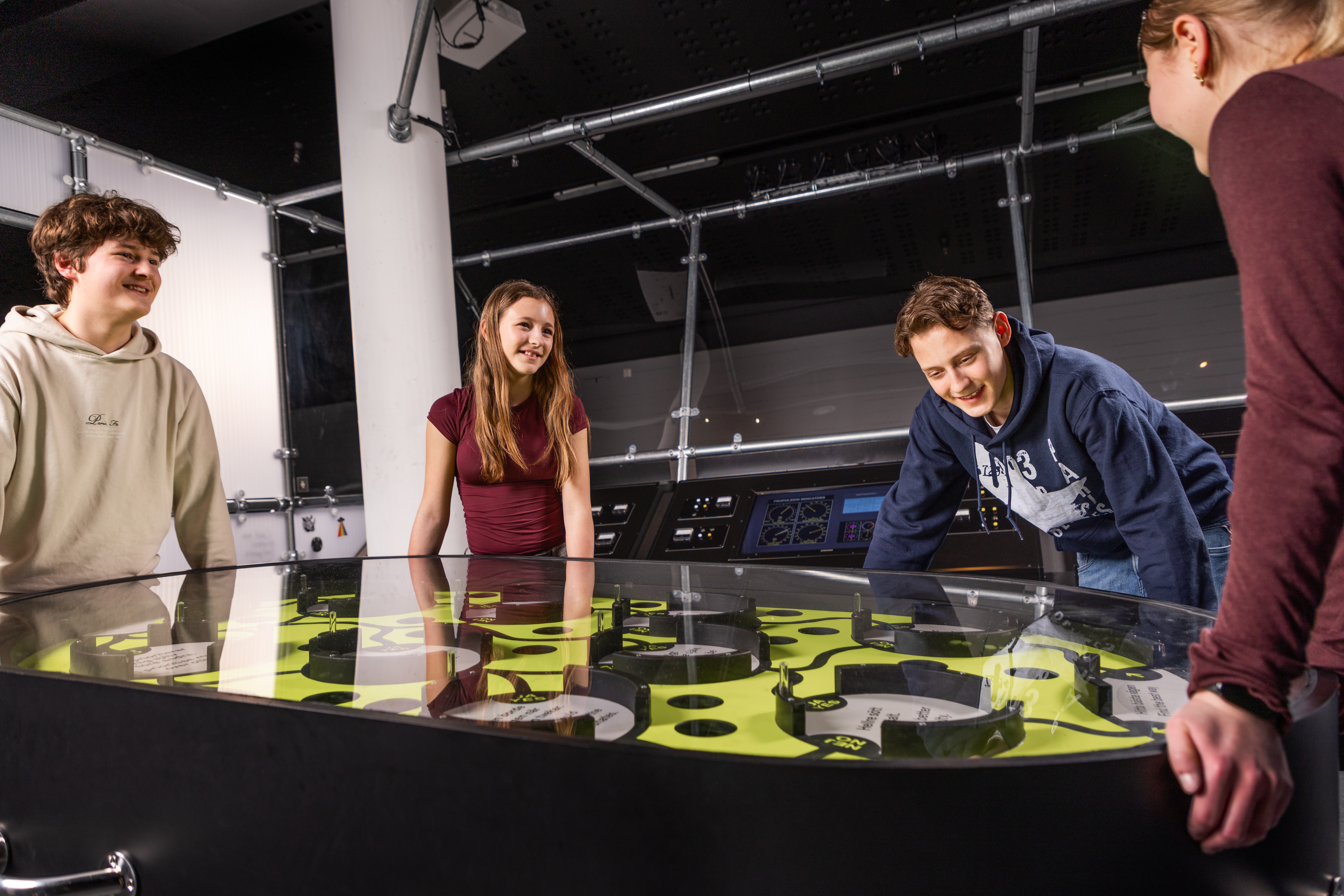 Four teenagers explore an interactive exhibit with a tabletop game in a modern museum setting.