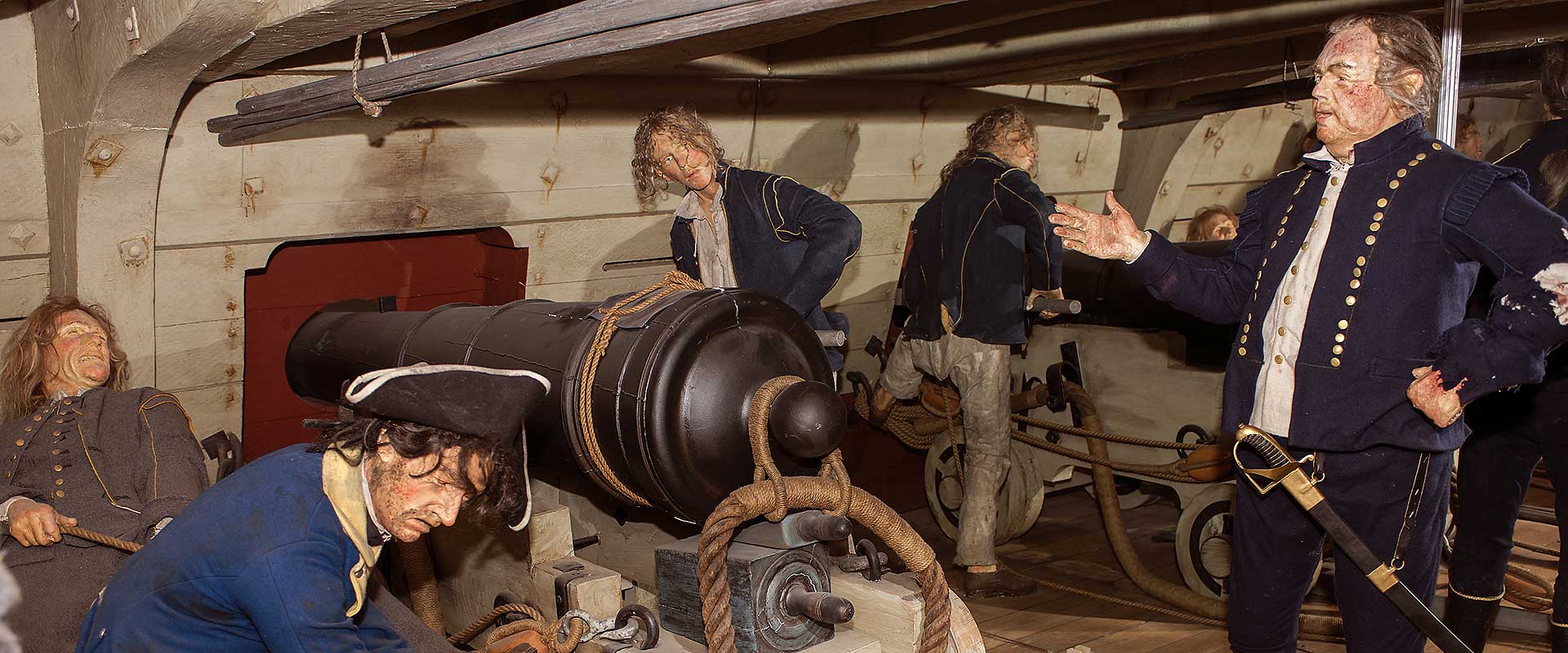 Diorama depicting the gun deck of an old naval warship, with realistic wax figures of sailors manning cannons during battle. An officer is giving orders while others load the guns or rest, exhausted.