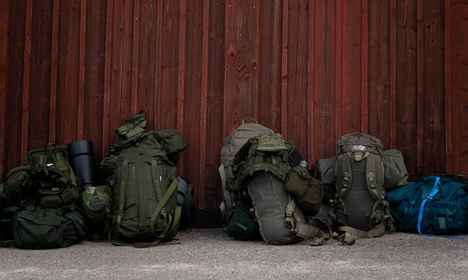 Four green backpacks are leaning against a red wooden wall.