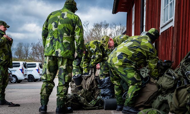 Several people in camouflage clothing stand taking off large backpacks by a red wall.