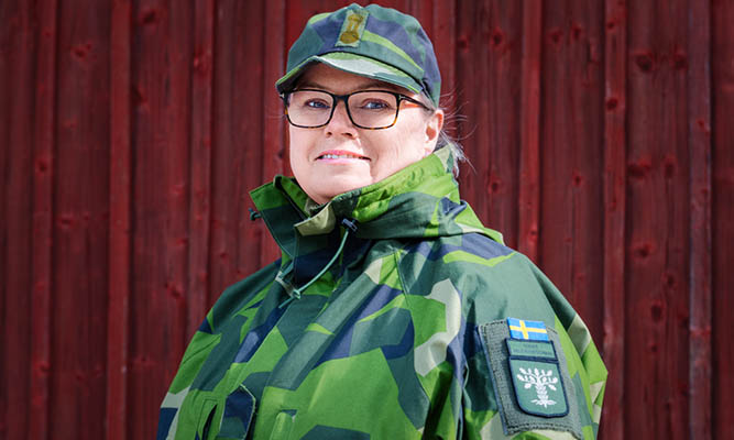 A woman in camouflage clothing stands in front of a red wooden wall and smiles into the camera.