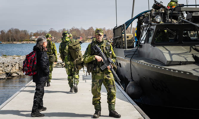 Men in camouflage clothing walk on a pier next to a military boat.