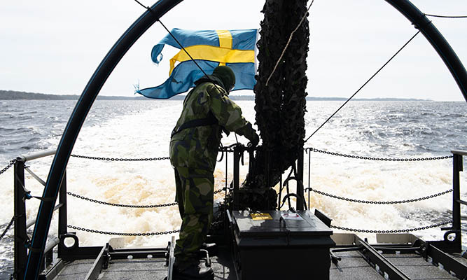 A person in camouflage uniform stands on a military boat with a Swedish flag flying at the stern, moving through open water.