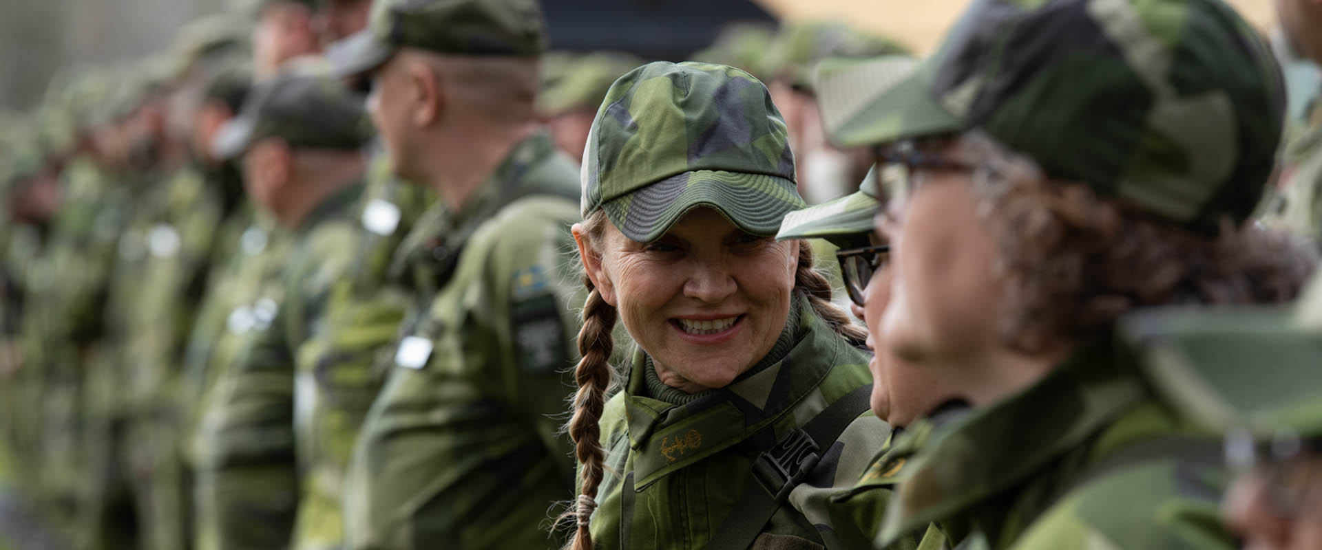 A group of people in camouflage uniforms stand in a line outdoors, with two women in the foreground smiling and talking to each other.