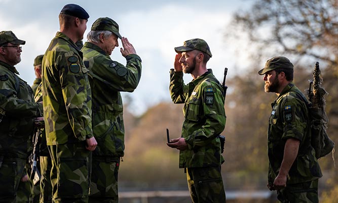 Four men in camouflage clothing stand opposite each other, two of them salute.