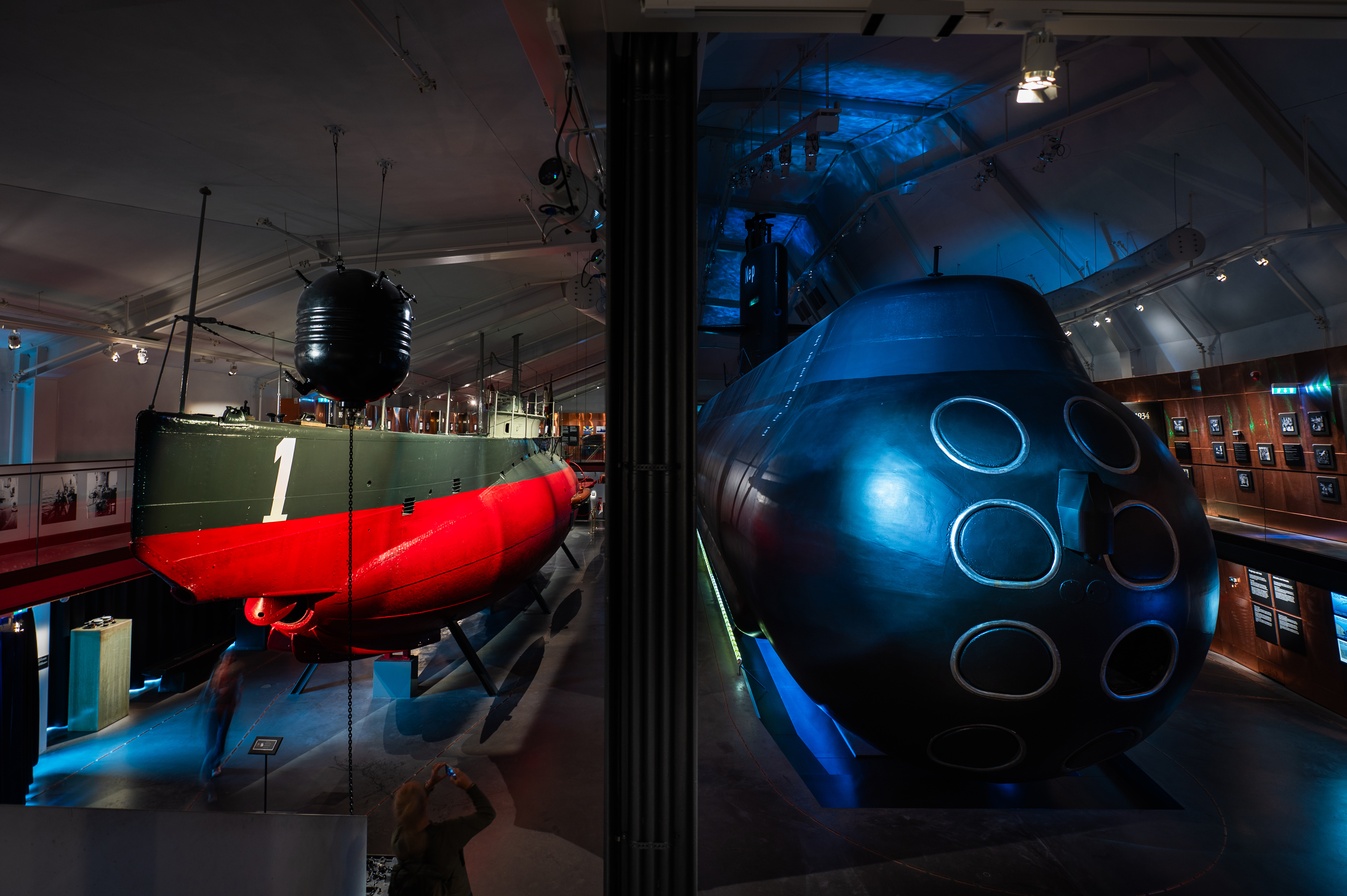 Two submarines on indoor display at a museum. On the left is an older, smaller submarine painted red and green with the number "1" on the bow and a naval mine suspended above. On the right is a larger, more modern submarine with multiple torpedo tubes on the front, illuminated with blue lighting. Visitors are seen observing and taking photos in the dimly lit exhibition space.