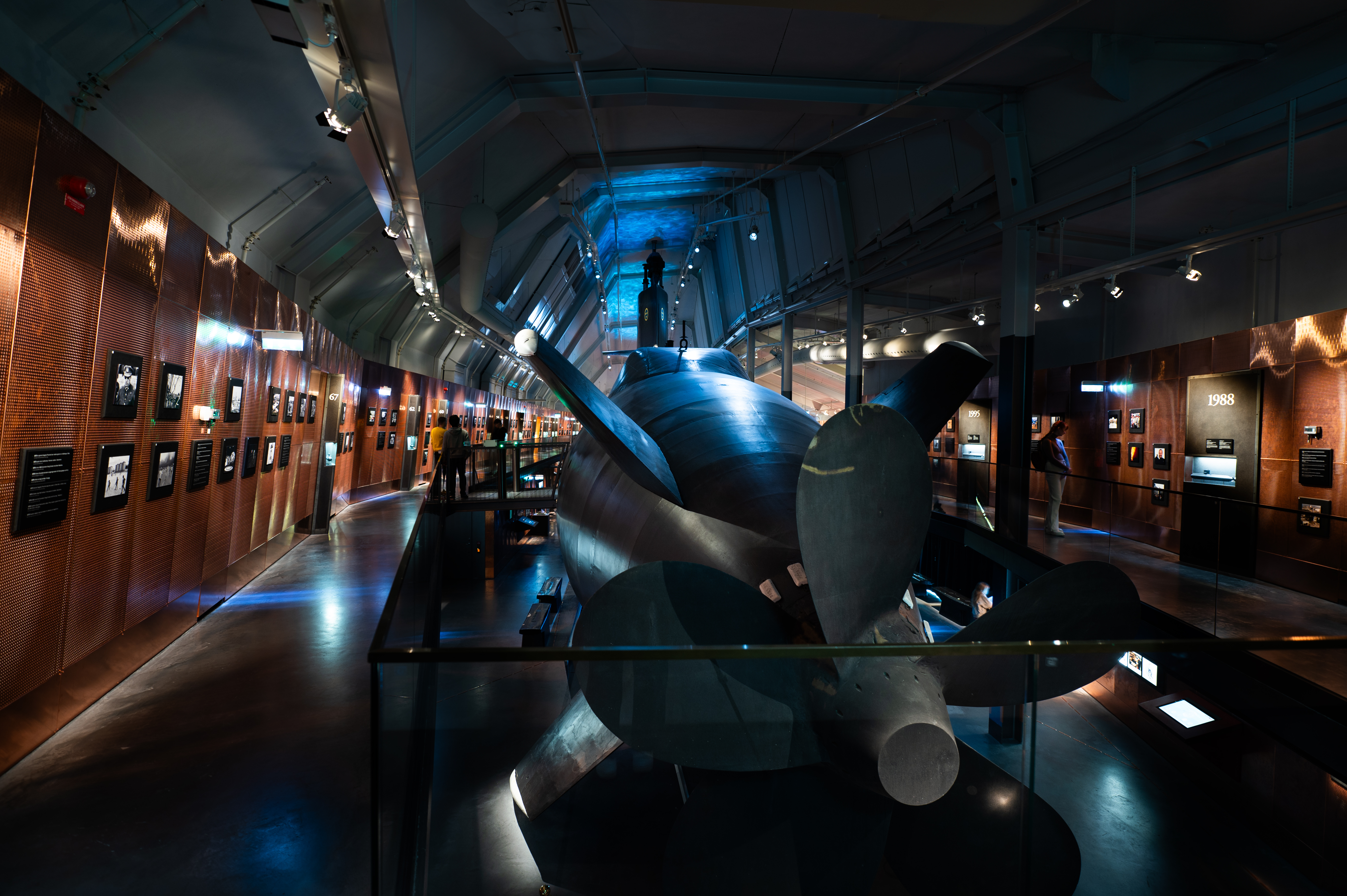 Rear view of a large submarine on indoor display at a museum. The image shows the submarine’s propeller and rudder, with a walkway around it where visitors are viewing exhibits along the walls. The lighting is dramatic, with blue and warm tones, and a wall display includes the year "1988".