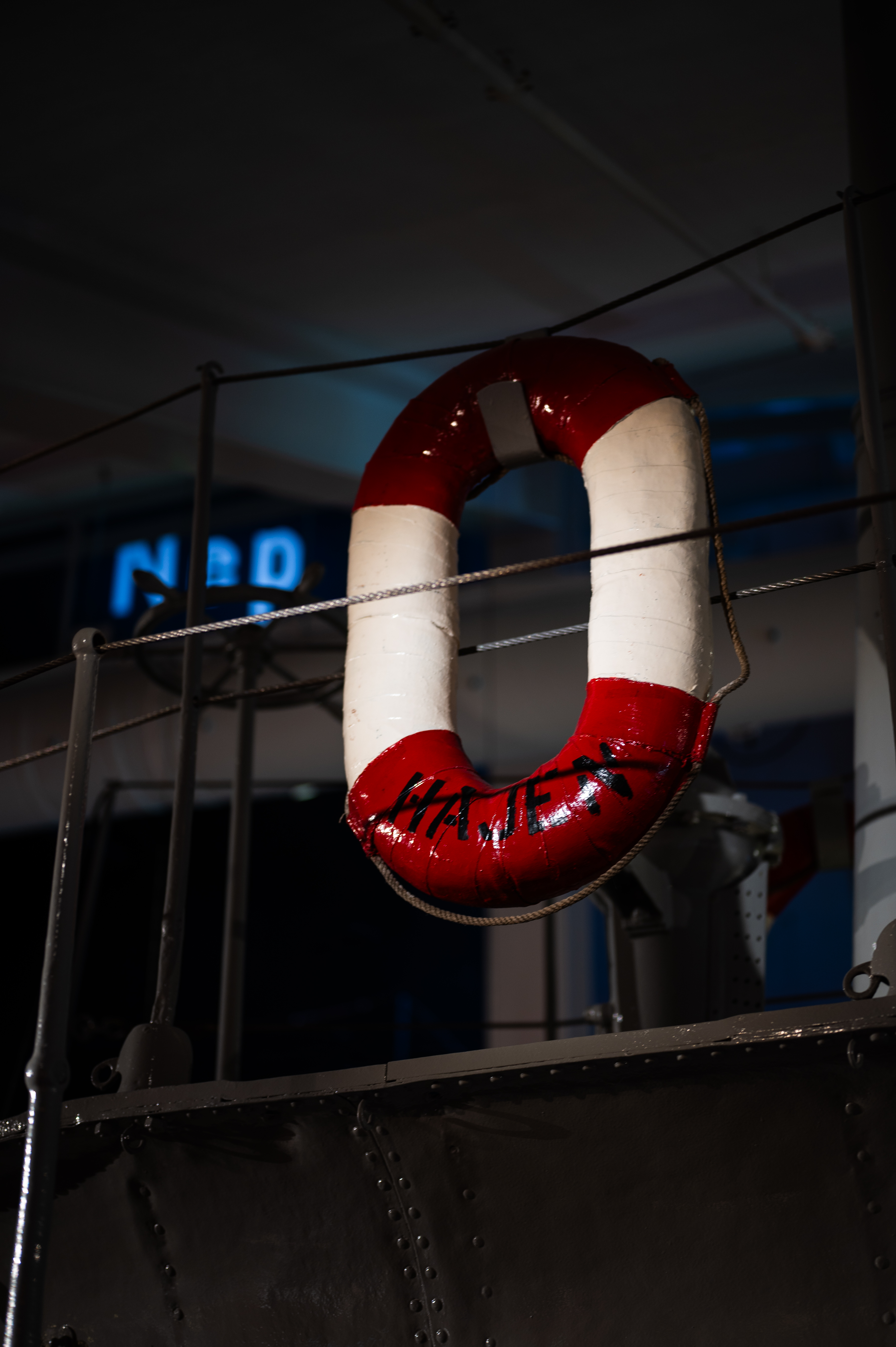 Close-up of a red and white lifebuoy with the text "HAJEN" mounted on a railing, indoors at a museum. The lifebuoy hangs by a rope, with part of the vehicle’s dark gray metal body visible in the background.