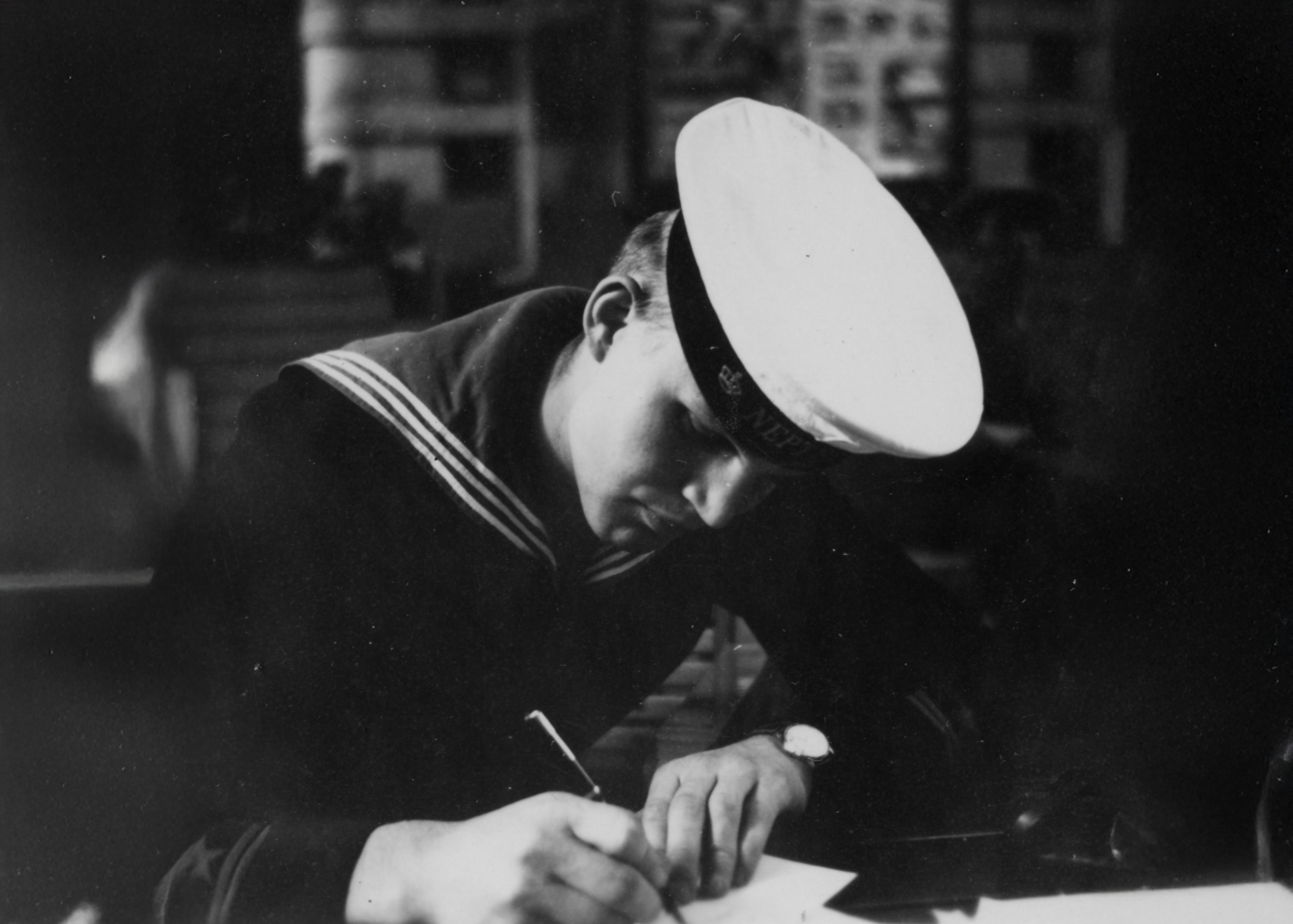 En svartvit bild som visar en matros i uniform med mössa märkt "HMS Neptun" som sitter och skriver ett brev vid ett bord.