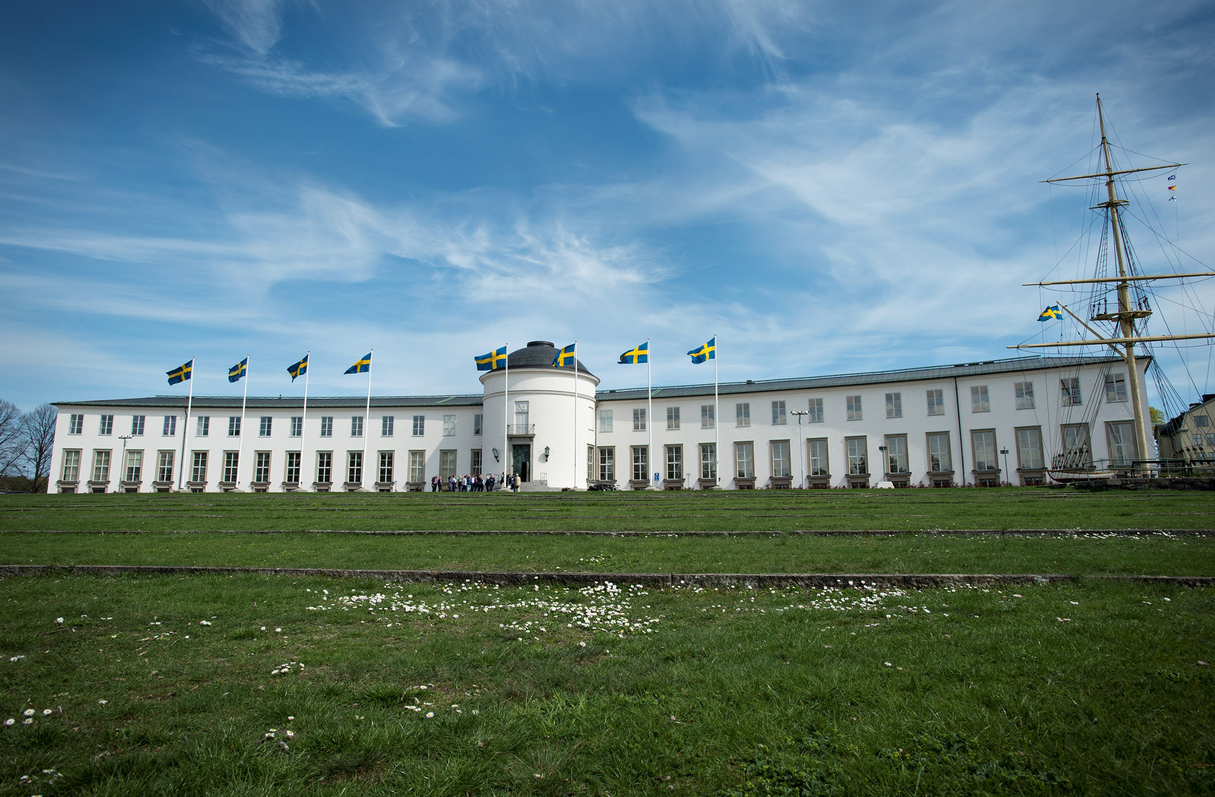 The white façade of a museum building seen from the front, with a lawn in the foreground. Swedish flags are waving in front of the building.