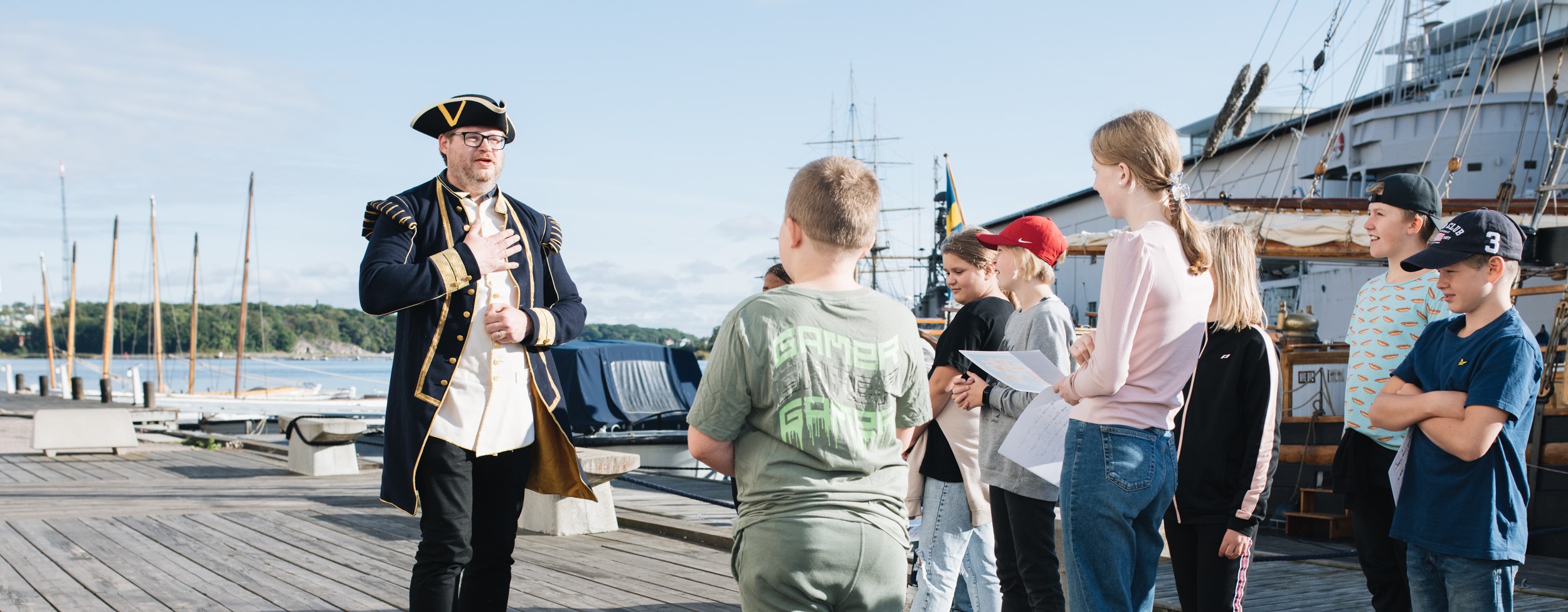 En man klädd i historisk uniform talar till en grupp barn på en kaj vid vattnet.