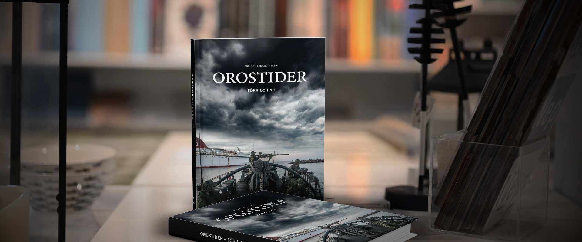 En bok med titeln Orostider – Förr och nu står upp på ett bord med ett exemplar liggande bredvid, mot en bakgrund av suddiga bokhyllor.