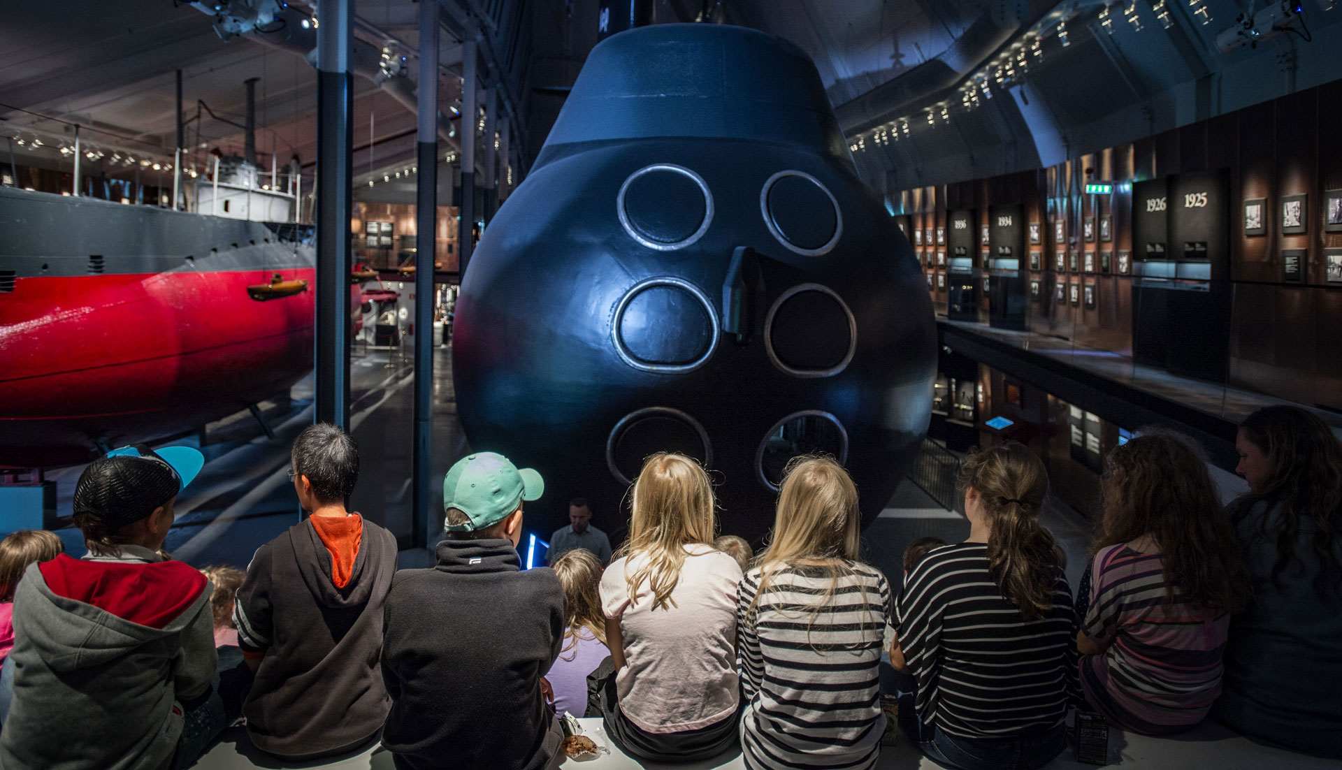 En grupp med barn sitter och lyssnar på en vuxen framför en ubåt i Ubåtshallen på Marinmuseum